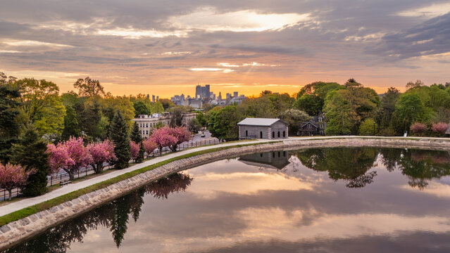 Aerial view of Brookline Reservoir with its stone gatehouse and blooming cherry blossoms against the distant Boston skyline at sunrise. Brookline, Massachusetts, United States.