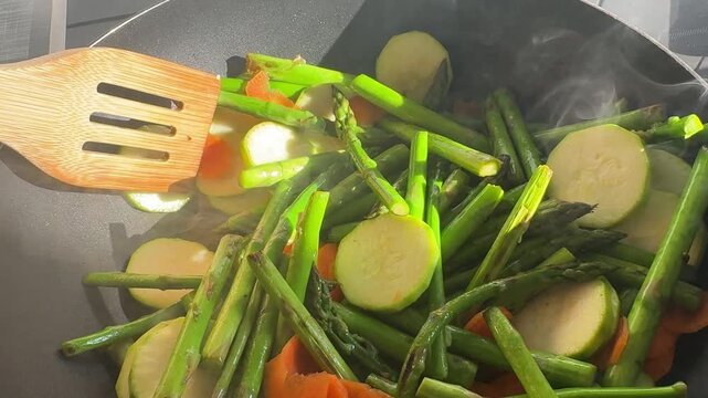 Wooden spatula stirring asparagus, zucchini and carrots in a steaming pan. Healthy vegetable stir fry and vegan cooking concept.