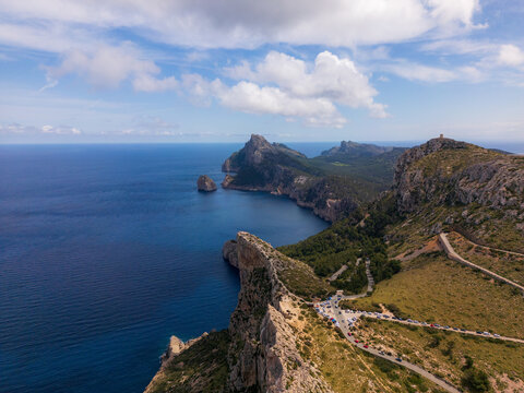 Aerial view of Cap de Formentor featuring rugged limestone cliffs, the deep blue Mediterranean Sea, and a winding coastal road with parked cars in Balearic Islands, Spain.