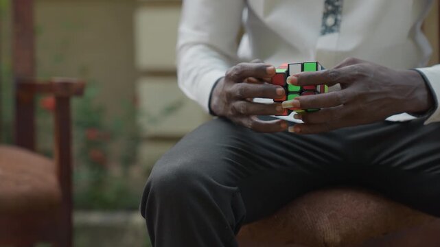 Closeup of black hands solving cube on lap, white shirt and patterned tie, seated on wooden chair in garden, slow deliberate twists, textured trousers and warm sunlight create intimate contemplative