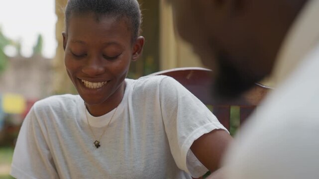 Smiling Black woman on shy date in sunlit courtyard, white tee and pendant, looking down with soft blush, playful laughter, receptive partner nearby, tender romantic moment, gentle eye contact, warmth