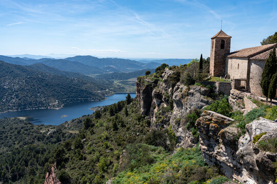 High-angle landscape of the historic stone church in Siurana perched on a limestone cliff overlooking the turquoise reservoir and the Prades Mountains under a clear blue sky.