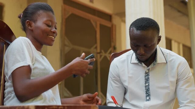 Black couple laughing during study date. Woman laughs and shows phone while man writes notes, joyful break, light teasing, casual porch setting, candid chemistry and warm smiles between partners