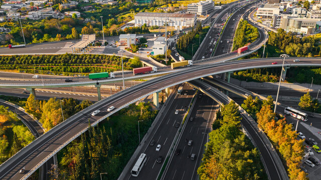 Aerial view showing a modern highway interchange with multiple elevated roads and ramps, cars, buses, and semi-trucks actively traveling, surrounded by green foliage and urban infrastructure