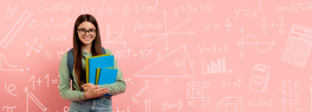 A student stands and smiles while holding several books. Behind her are various math and science illustrations on a pink wall. This scene takes place in a classroom during the school day.