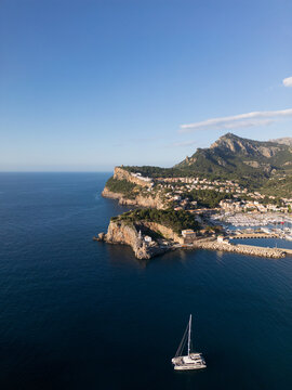 Aerial view of the harbor with a lighthouse on the rocky cliffs and a catamaran sailing on the blue Mediterranean sea in Port de Soller, Balearic Islands, Spain.