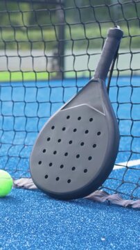 Vertical video: Camera zooming to black padel paddle on court with green ball and net, showing grip