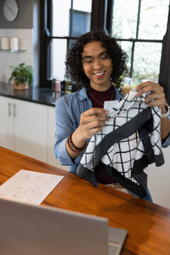 Non-binary adult inspecting grid garment tag at wooden kitchen island with laptop and sketches