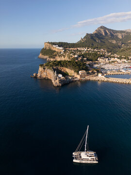 Aerial view of Port de Soller with a lighthouse on a rocky cliff and a white catamaran sailing on the deep blue sea Port de Soller, Balearic Islands, Spain.