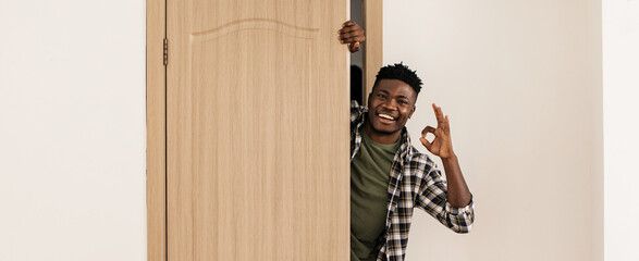 A man stands at a wooden door smiling and making an OK gesture with his hand. He appears to be in a bright and simple room. The door is open slightly, revealing a light-colored wall. © Prostock-studio
