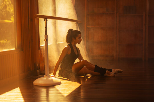 Ballerina in black sportswear practicing at a barre in a sunlit wooden studio.