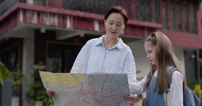Portrait of happy mother holds map and girl points at landmarks near old buildings. Woman with little daughter explores famous sightseeings on holiday