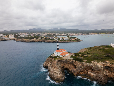 Aerial view of Far de Portocolom lighthouse on a rocky cliff overlooking the Mediterranean Sea with the town and mountains in the background Portocolom, Balearic Islands, Spain.