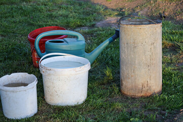 Various garden tools, including a plastic watering can and several buckets filled with water, stand on green grass during evening chores. An old metal bucket and bright plastic containers are ready fo © Олег Струс