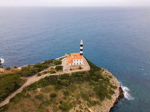 Aerial view of the Far de Portocolom lighthouse with its iconic black and white stripes on a rocky cliff overlooking the blue sea Portocolom, Balearic Islands, Spain.