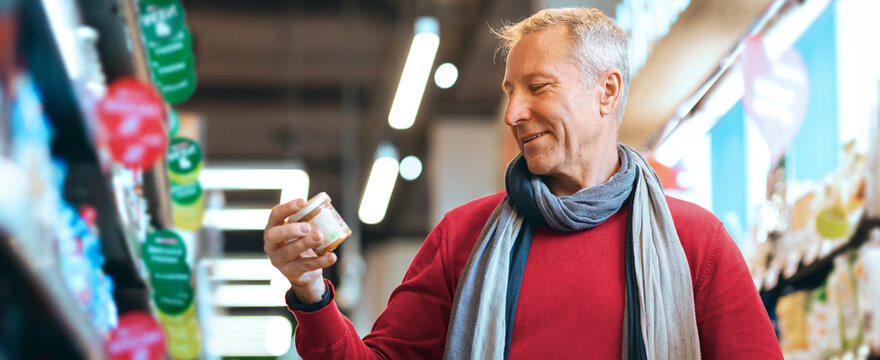 A man is holding a jar while looking at it in a grocery store. He appears to be checking the label. Shelves are filled with various food items. Bright lights are overhead.
