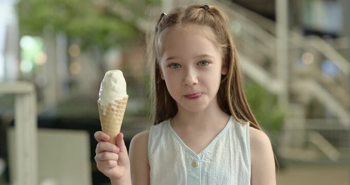 Portrait of little girl licks lips as holds melting ice cream in summer urban park. Cute child with pigtails eats delicious sundae in wafer cone on city street