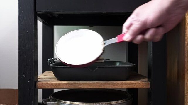 Close-up view of a person taking a small, white frying pan from a wooden shelf in a kitchen cabinet to prepare for cooking a meal