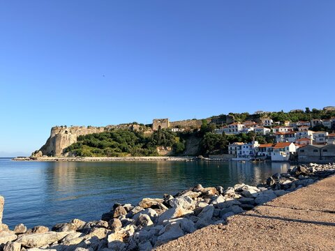 Koroni, Greece, Coastal town with historic stone fortress and red roof houses by calm sea under clear blue sky