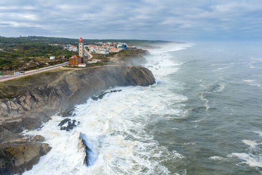 Aerial view of the red-topped Farol Penedo da Saudade lighthouse on rugged cliffs with crashing waves and coastal town under a cloudy sky in Marinha Grande, Leiria, Portugal.