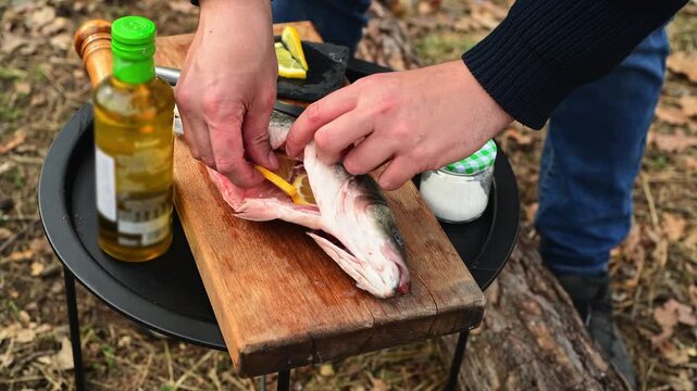Close up of male hands stuffing fish with lemon slices before cooking over a campfire. Outdoor food preparation scene in the forest with camping atmosphere.