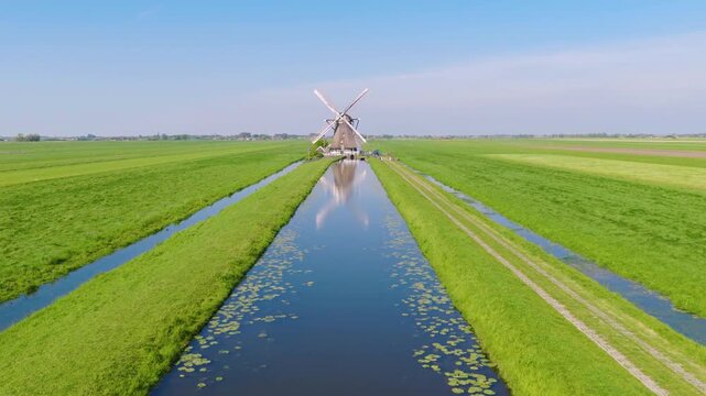 Aerial view of traditional Dutch windmills near water canals in a green field. Clear sunny day with reflections on the water surface and a wide video landscape captured by drone.