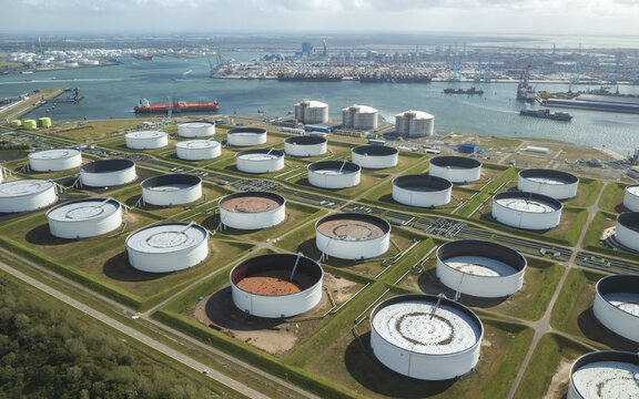 Aerial view of numerous white circular oil storage tanks in the Port of Rotterdam, Netherlands, arranged in a grid near a busy industrial harbor with cargo ships and cranes under a bright sky.