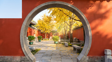 Obraz premium Autumn Chinese garden gate, Circular moon doorway with yellow foliage, Traditional red wall and stone path, Serene courtyard travel scene