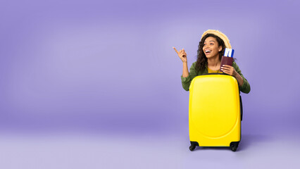 A woman stands next to a yellow suitcase while holding travel documents and passport. She is smiling and pointing, showing her excitement for the upcoming journey. © Prostock-studio