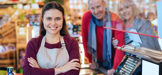 A shopkeeper stands at the register with a smile while customers look on. The store is busy with colorful displays of products in the background during the afternoon. © Prostock-studio