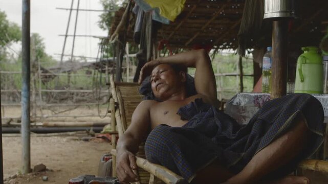 Tired shirtless worker resting on a bamboo chair after hard work in a makeshift camp in Myanmar