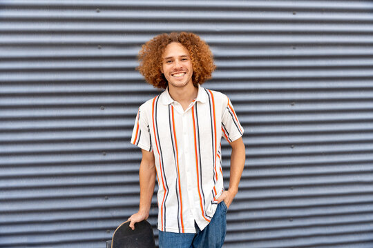 Smiling urban skater holding skateboard by metal wall