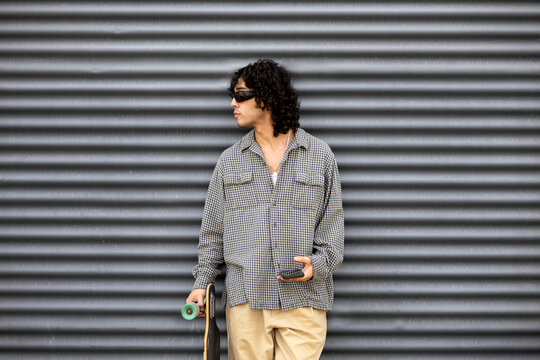 Urban skater holding long board by corrugated metal wall