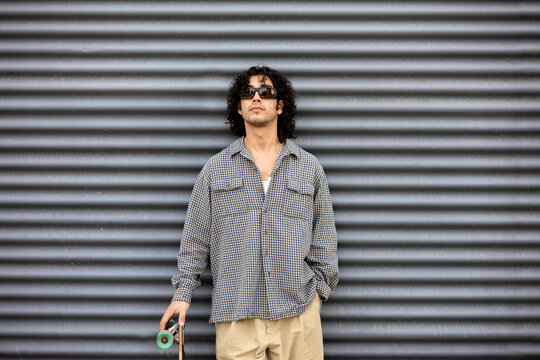 Young man with skateboard in sunglasses by metal wall