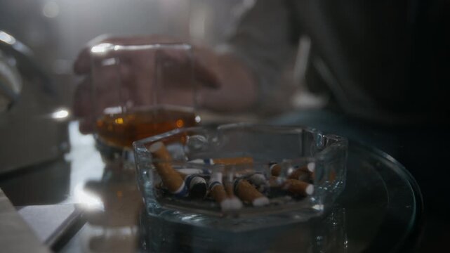 Close up view of hands of unrecognizable man stubbing cigarette into glass ashtray filled with butts and then drinking whiskey from glass at table