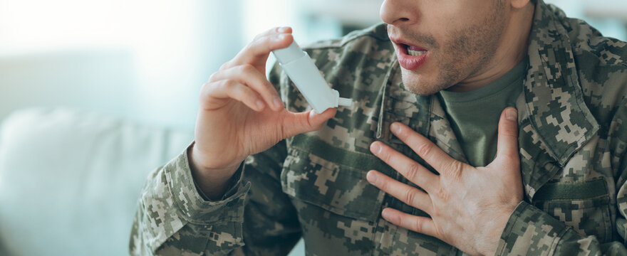 A man in military uniform holds an inhaler while sitting on a couch at home. He appears to be having difficulty breathing and is using the inhaler for relief.