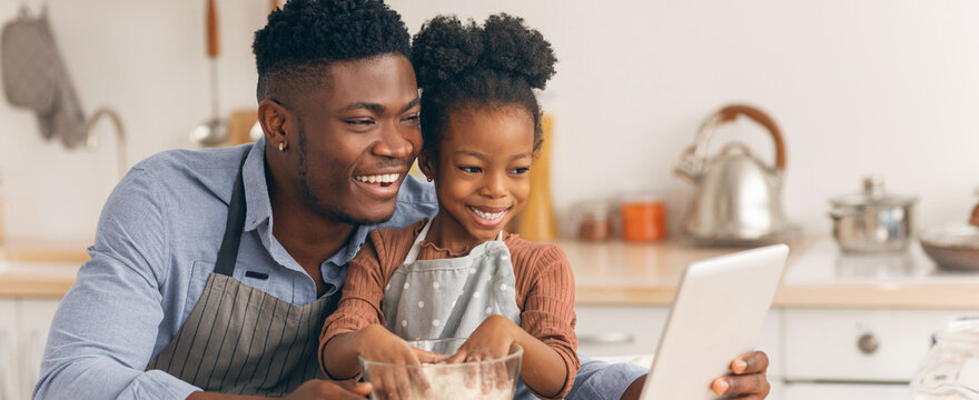 A father and daughter are preparing food in their kitchen. They are mixing ingredients in a bowl while following a recipe on a tablet. They enjoy their time together.