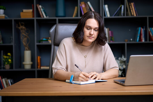 Woman writing ideas in notebook while working on laptop at desk