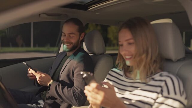 Smiling bearded man in blazer holding smartphone, sitting in driver&rsquo;s seat and looking at woman in striped shirt beside him. Both appear cheerful, sharing moment inside parked car on sunny day.