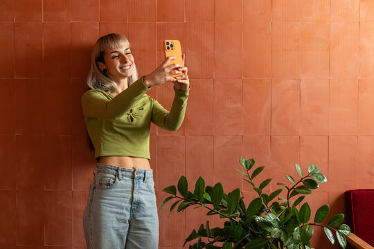 Smiling young woman taking smartphone selfie by wall