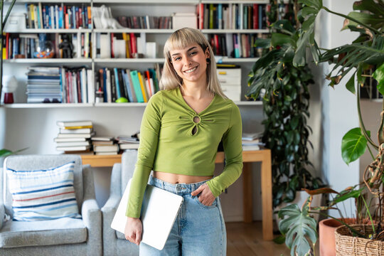 Portrait of smiling woman holding laptop at home office bookshelf
