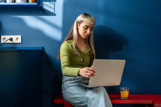 Woman using laptop on bench in blue kitchen with sunlight