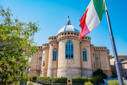 Pontifical Basilica della Santa Casa in Loreto framed by its dome and red tile roofs, Italian flag waving against a clear blue sky historic pilgrimage landmark in Marche, Italy