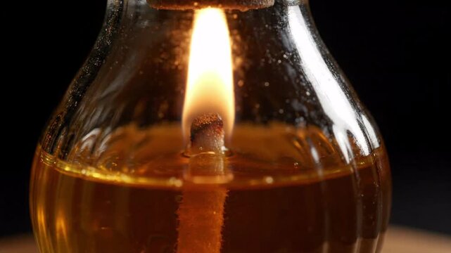 Close up of an oil lamp wick burning with a bright yellow and orange flame inside a glass container filled with golden liquid, warm glowing light on dark background
