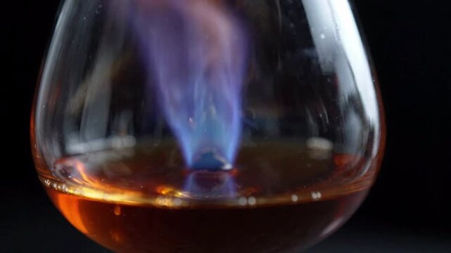 Flamb? technique, blue flames burning in a brandy snifter glass with amber liquid, dark background, close-up macro shot, culinary preparation, alcohol vaporization