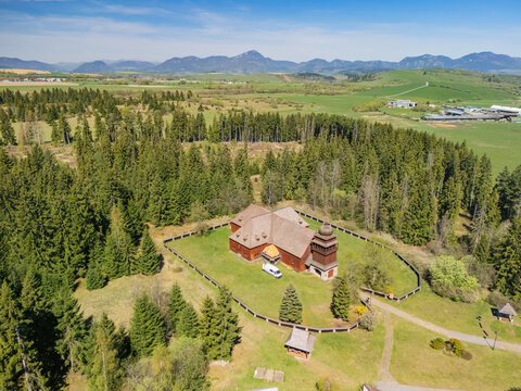 Aerial view of the Wooden Articular Church of Svaty Kriz surrounded by lush evergreen forests and rolling hills under a clear blue sky in Svaty Kriz, Zilinsky kraj, Slovakia.