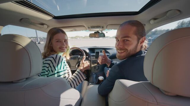 Caucasian woman and bearded man sitting in car front seats turning heads back at camera. Male and female passengers posing inside modern vehicle and giving thumb up in daylight during travel.