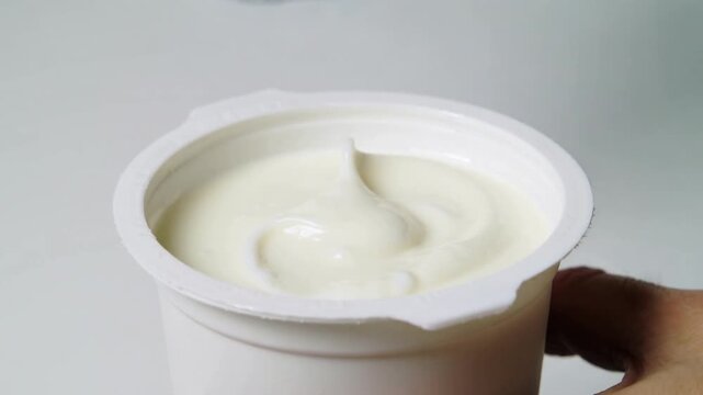 Pinching foil tab, adult hands pulling back foil lid at tabletop, exposing creamy yogurt inside cup