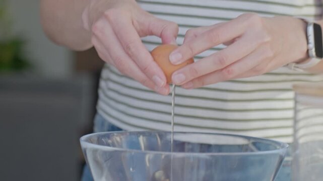 Woman cracks egg and adds it into glass bowl on kitchen counter