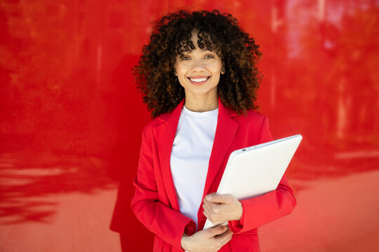 Confident Hispanic woman in red suit holding laptop